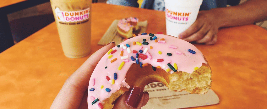 A hand holds a pink and white frosted doughnut, with two drinks in the background; one of the drinks is the the Dunkin' Donuts birthday reward.