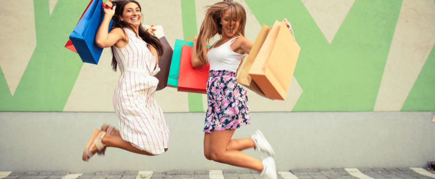 Two women joyfully jump while holding colorful shopping bags filled with fashion birthday freebies.