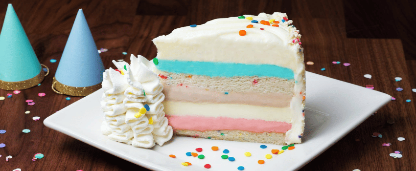 A slice of colorful layered cake with sprinkles and whipped cream on a white plate, surrounded by party hats and confetti. The dessert is The Cheesecake Factory birthday reward.